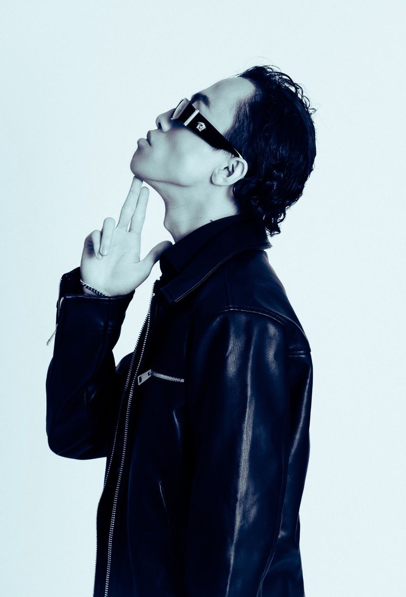 Man in side profile wearing dark sunglasses and a black leather jacket with fingers under his chin against a plain backdrop.