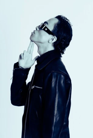 Man in side profile wearing dark sunglasses and a black leather jacket with fingers under his chin against a plain backdrop.