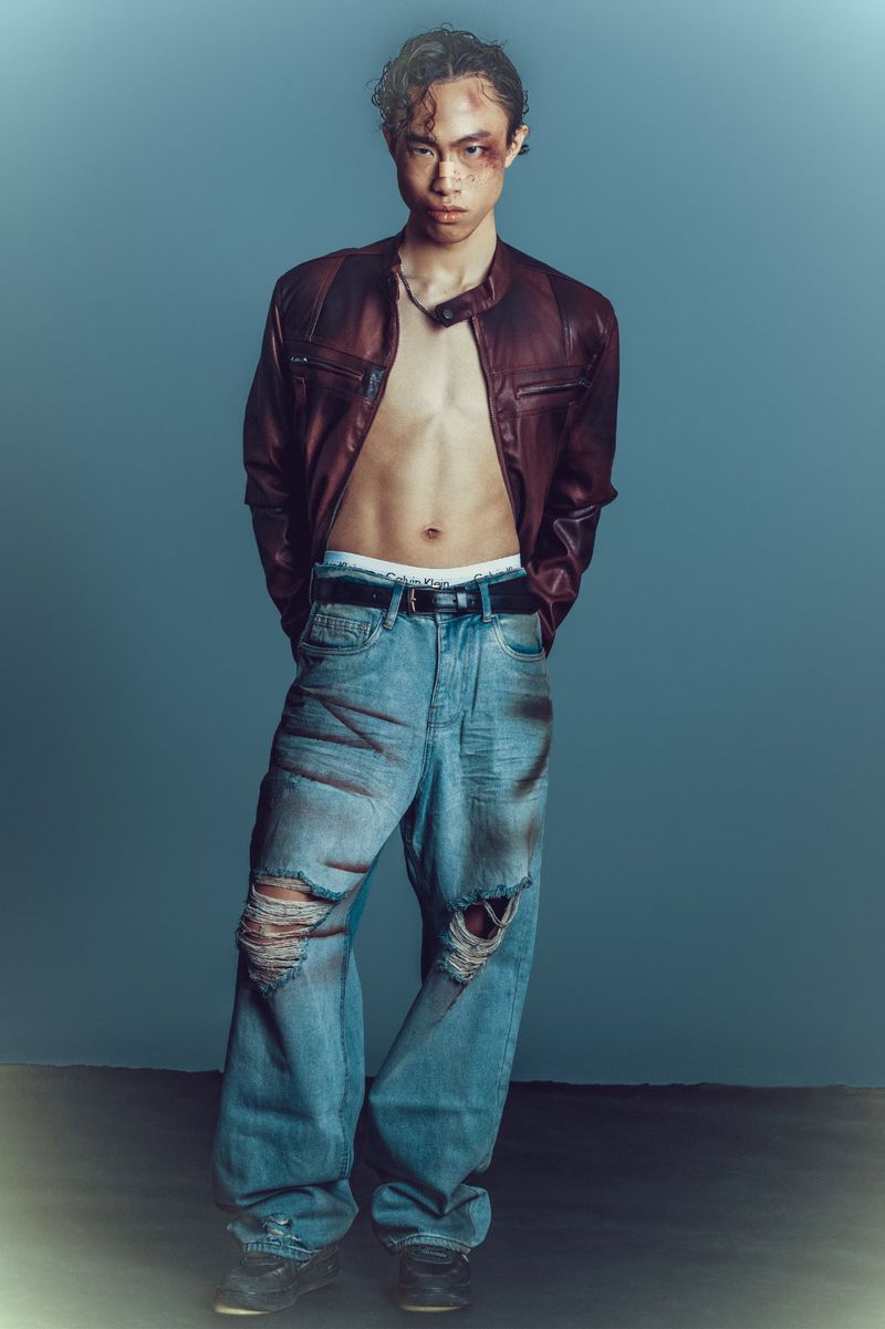 Full body studio portrait of an Asian male wearing a maroon leather jacket and ripped jeans with a mark around one eye looking at camera.