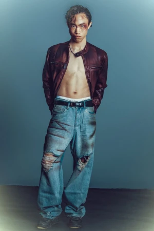 Full body studio portrait of an Asian male wearing a maroon leather jacket and ripped jeans with a mark around one eye looking at camera.