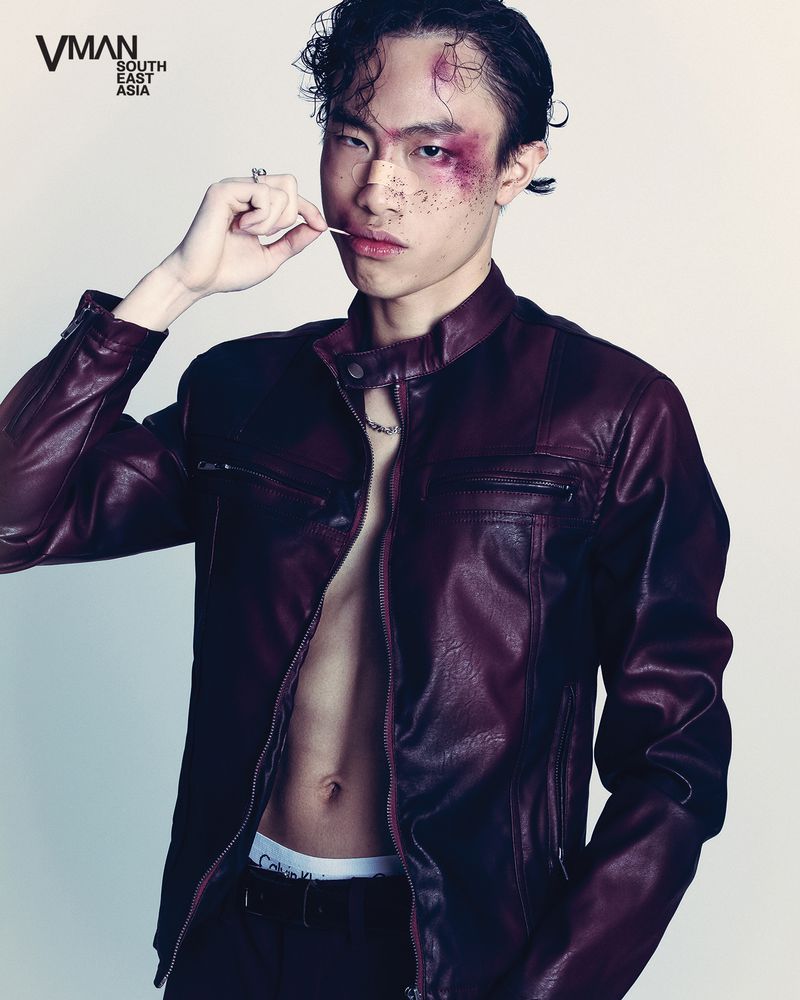 Portrait of a young Asian man with a faux facial injury, wearing a burgundy leather jacket and lip jewelry, against a white background.