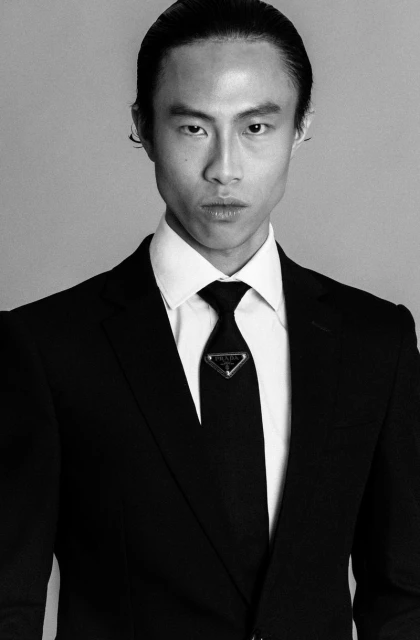 Portrait of an adult Asian male wearing a black suit with a white shirt and black tie, standing indoors with a neutral expression.