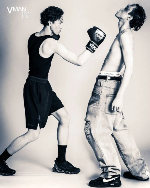 Two young adult men in a boxing pose, one wearing boxing gloves, tank top, and shorts, the other shirtless with jeans, in a studio setting.
