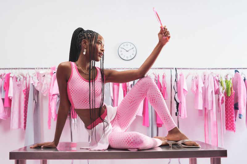 Woman in pink activewear set posing on a metal table holding a hand mirror in front of a clothing rack filled with pink apparel.