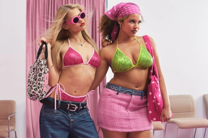 Two models pose in sequined bikini tops, low-rise denim jeans, and a pink mini skirt in front of a pink curtain backdrop.
