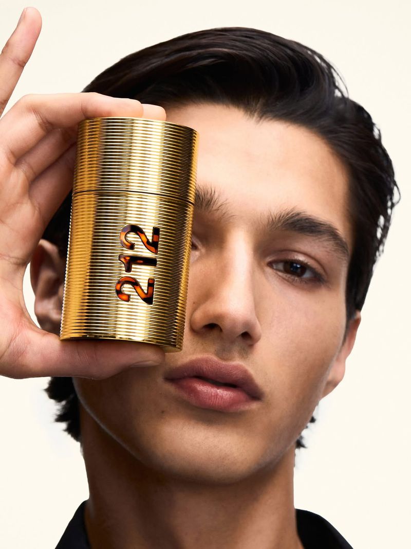 A male model holding a ribbed gold 212 Men Parfum bottle in front of his eye against an off-white background.