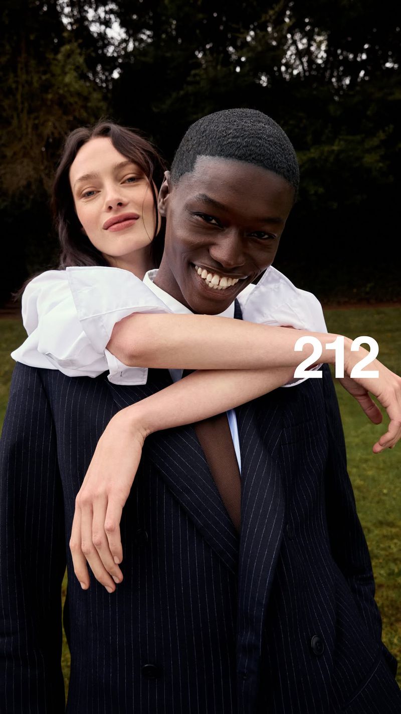 Man in a navy suit carrying a smiling woman on his back outdoors near a park background with white 212 text overlay.