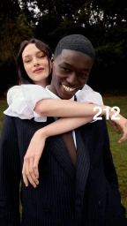 Man in a navy suit carrying a smiling woman on his back outdoors near a park background with white 212 text overlay.