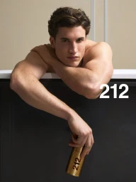Shirtless man leaning over a dark ledge holding a gold 212 perfume bottle against a neutral beige wall.