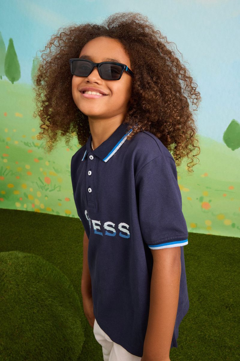 Young boy with curly hair smiling in black sunglasses and a navy blue Guess polo shirt in a set-dressed studio.