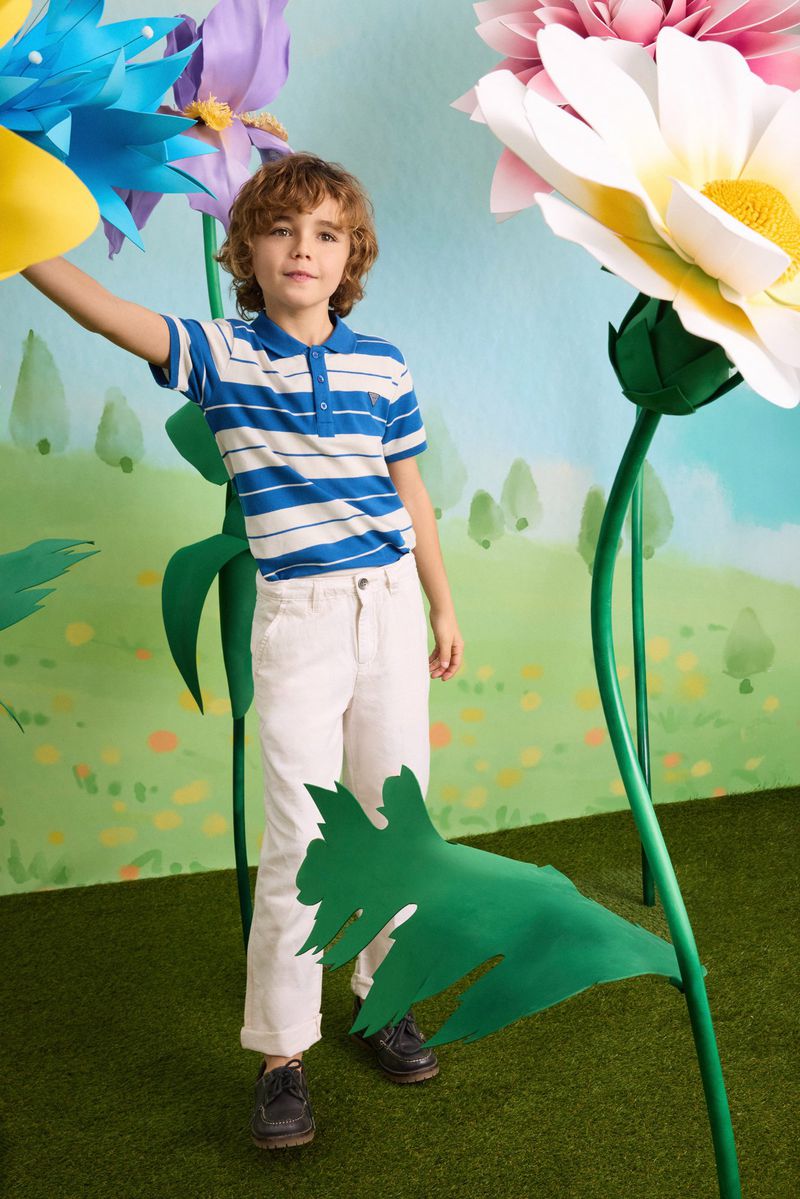 Young boy in a blue and white striped polo shirt and white trousers posing beside giant colorful paper flowers on a set.