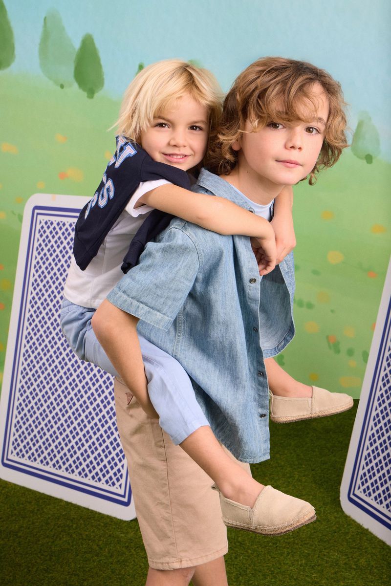 Two smiling young boys in summer casual wear, one giving the other a piggyback ride against an illustrated storybook backdrop.