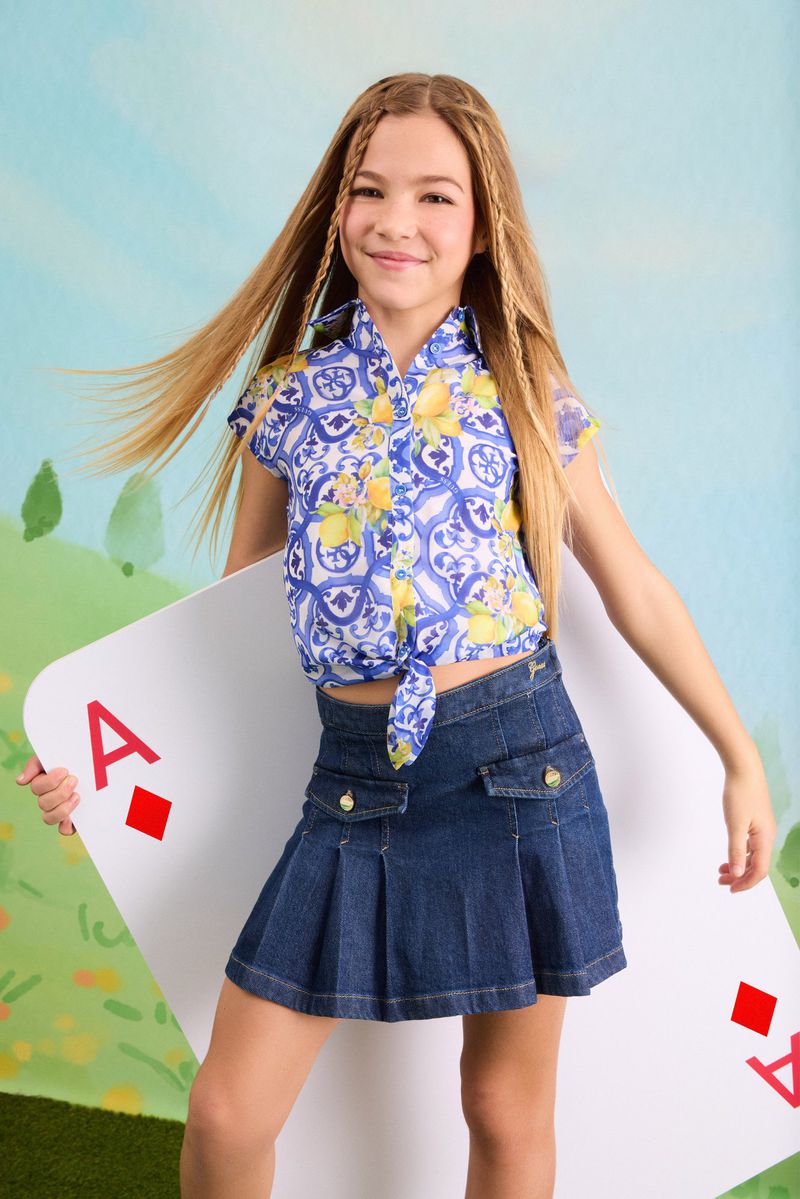 Young girl posing in a blue and white lemon print tie shirt and pleated denim skirt holding a giant ace of diamonds card.