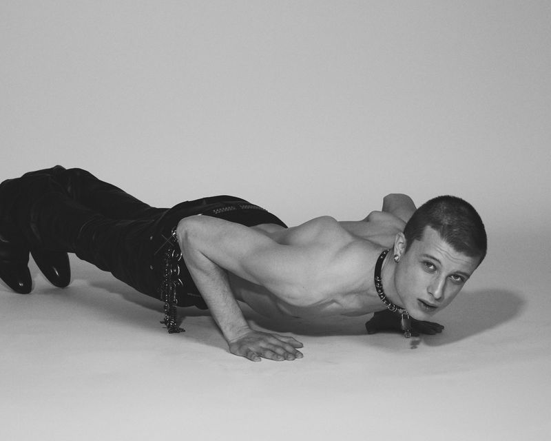 Shirtless young man in black leather pants and studded choker holding a low pushup pose against a seamless studio backdrop