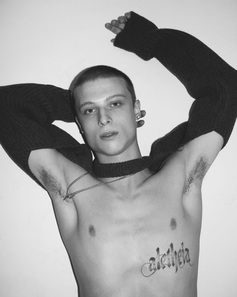 Shirtless young man with buzz cut and chest tattoo posing with raised arms wearing black knit sleeve scarf against plain background.