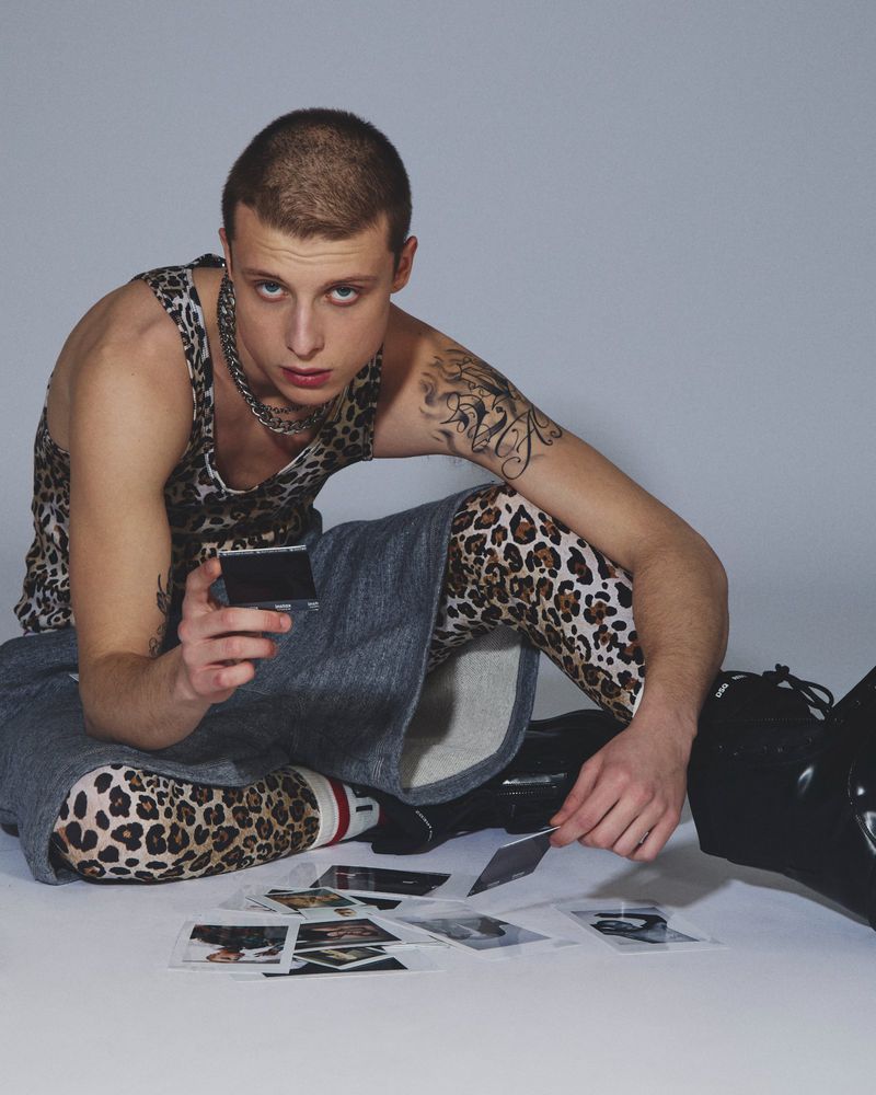 Man with buzz cut crouching in leopard print tank and tights looking at instant photos on a studio floor.