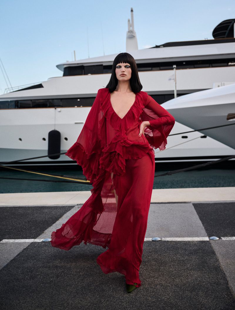 Model Daphne Groeneveld wearing a flowing sheer red ruffled gown on a marina dock in front of a white luxury yacht.