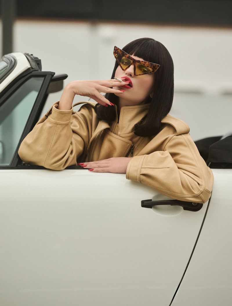 Model Daphne Groeneveld in tortoiseshell sunglasses and a camel jacket leaning against the door of a white convertible car.