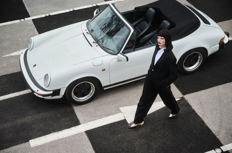 Daphne Groeneveld in a black pantsuit walking past a white convertible sports car on a checkered asphalt surface.