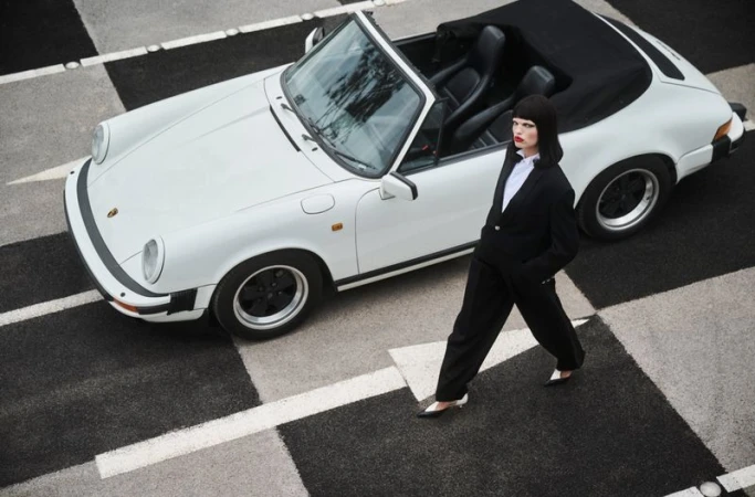 Daphne Groeneveld in a black pantsuit walking past a white convertible sports car on a checkered asphalt surface.