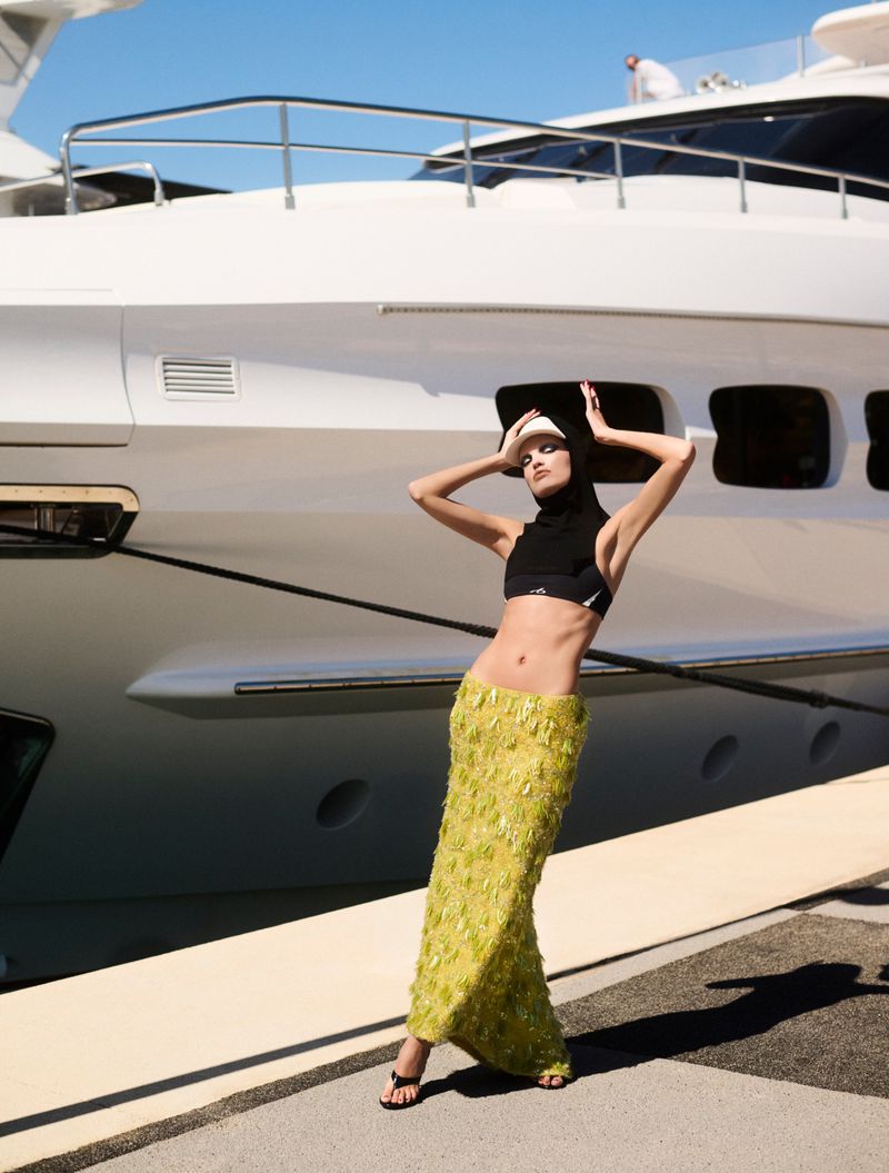 Model Daphne Groeneveld posing in a lime green textured maxi skirt and black hooded crop top before a luxury yacht on a marina dock.