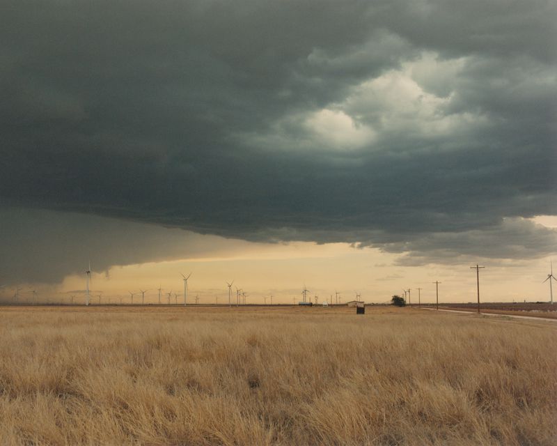 Dark storm clouds gather over a vast dry grass field with wind turbines and utility poles along the horizon.