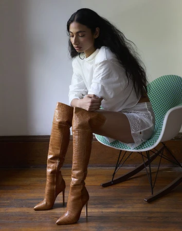 Woman seated in a green rocking chair wearing a white top, lace shorts, and tan croc-embossed thigh-high stiletto boots.