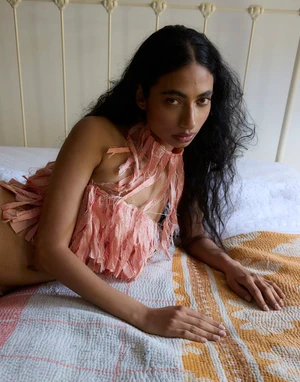 South Asian woman wearing a textured peach fringe halter top lying on a bed with a patterned quilt in front of a white headboard.
