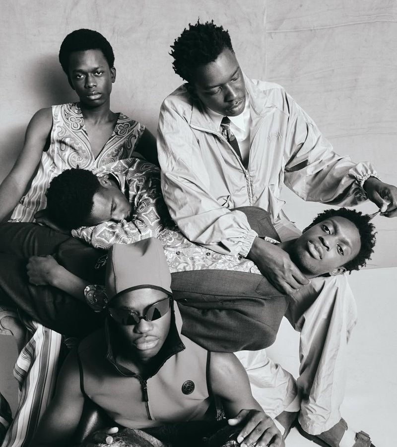Black and white studio shot of five African American men wearing vests, a jacket, and sunglasses, and their hair styled in afros.