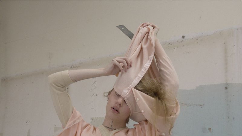 Model Wolfgang Beck pulling a pale pink buttoned garment over their head against a pale distressed studio wall.