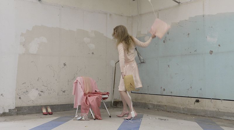 Model with long blonde hair wearing a pale pink outfit and heels, swinging a pink pillow in a distressed room with worn walls.