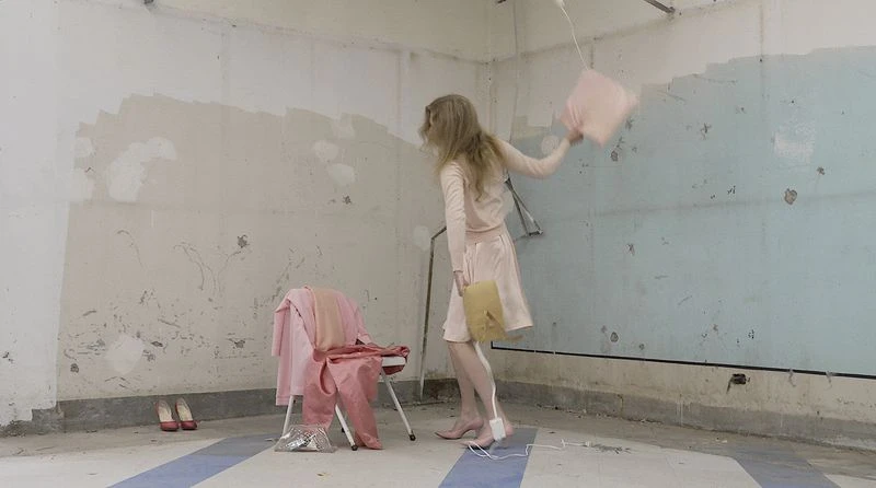 Model with long blonde hair wearing a pale pink outfit and heels, swinging a pink pillow in a distressed room with worn walls.