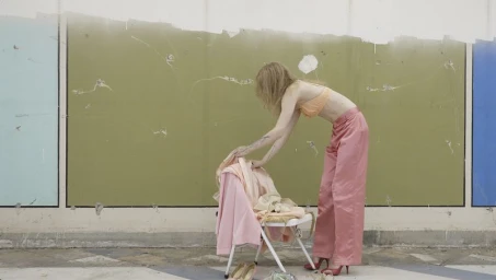Model bending over a folding chair holding pink garments in front of a weathered green wall.