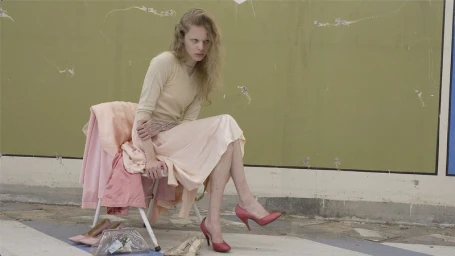 Wolfgang Beck seated on a folding chair wearing a cream top, pastel pink skirt, and rose heels against an olive green wall.