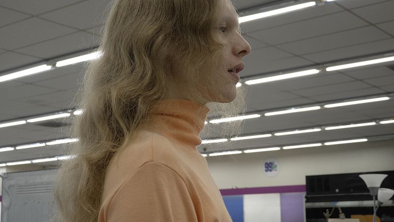 Model Wolfgang Beck shown in profile wearing a light peach turtleneck under fluorescent lights inside a retail store.