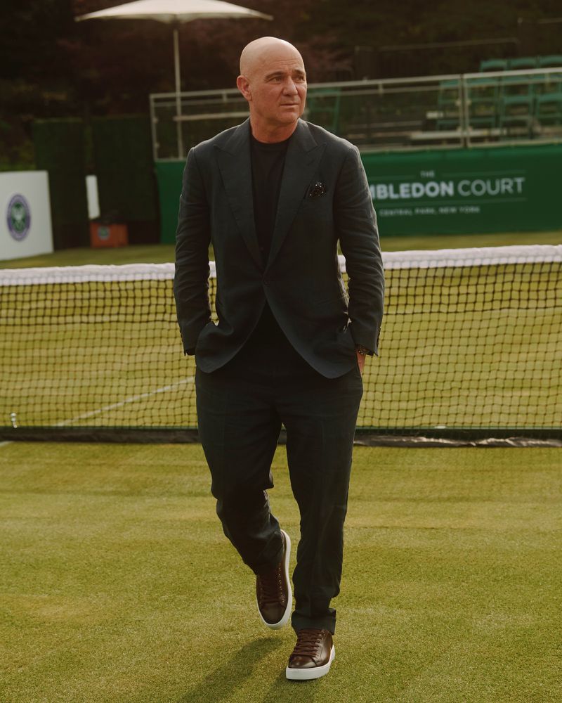 Andre Agassi in a dark tailored suit and sneakers walking across a grass tennis court with a tennis net in the background.