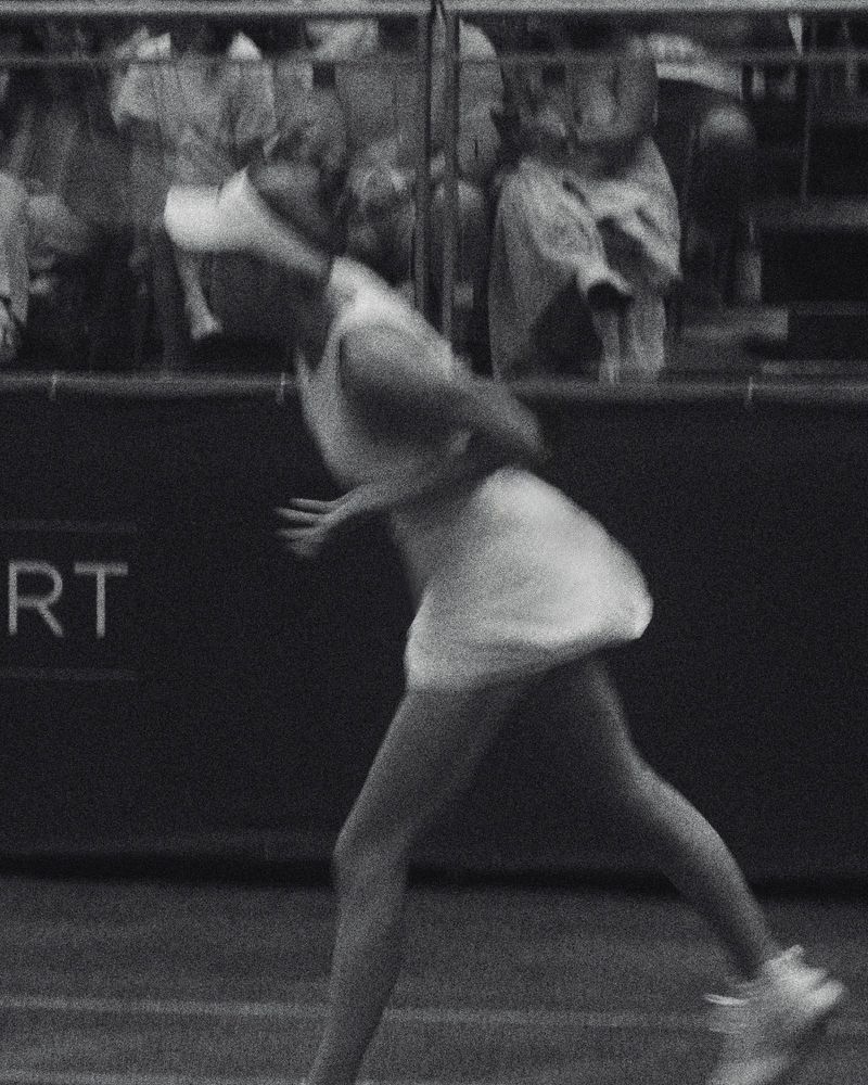 Black and white photograph of a tennis player in motion wearing a white visor and dress with spectators in the background.