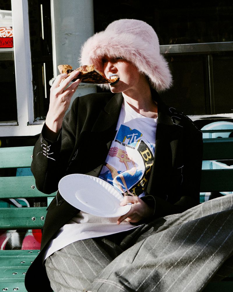 Model wearing a pink fuzzy bucket hat and black jacket eating a slice of pizza while seated outdoors on a green bench.