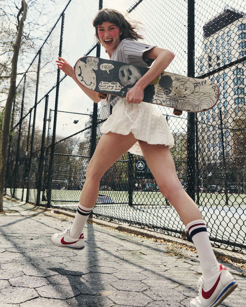 Model Lulu Röhr running outdoors holding an illustrated skateboard along a chain-link fence in a white skirt and sneakers.