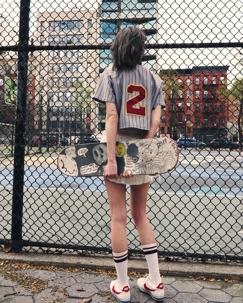 Woman viewed from behind holding an illustrated skateboard across her legs, wearing a cropped baseball jersey by a chainlink fence.