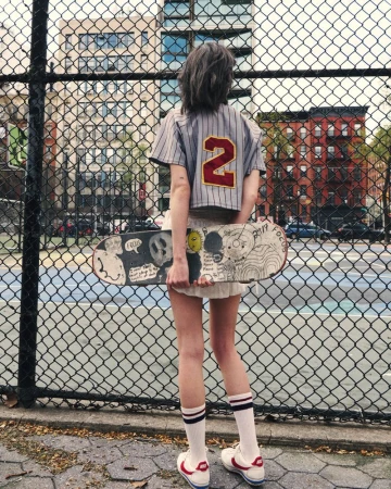 Woman viewed from behind holding an illustrated skateboard across her legs, wearing a cropped baseball jersey by a chainlink fence.