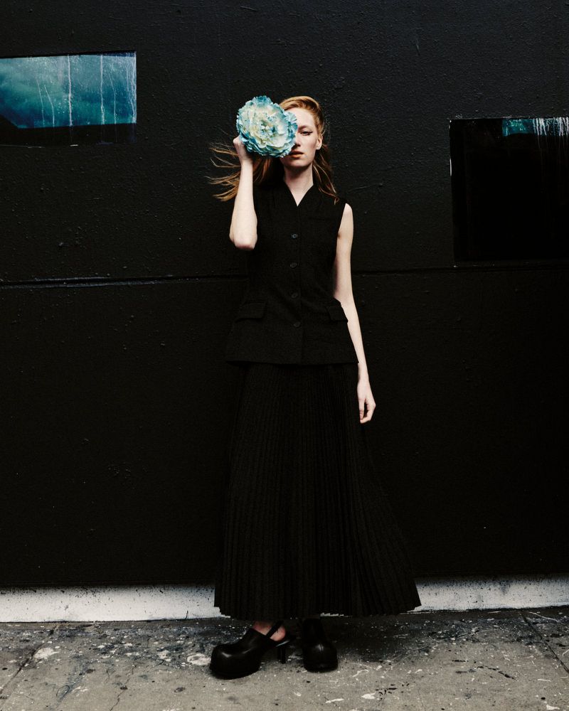 Model Lulu Röhr holding a blue cabbage over one eye while posing in a black vest and pleated maxi skirt against a dark wall.