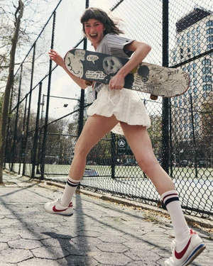 Model Lulu Röhr running outdoors holding an illustrated skateboard along a chain-link fence in a white skirt and sneakers.