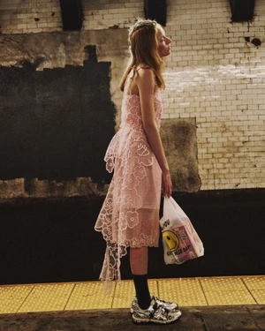 Model Lulu Röhr in a sheer pink floral lace dress and silver sneakers holding a plastic grocery bag on a tiled subway platform.