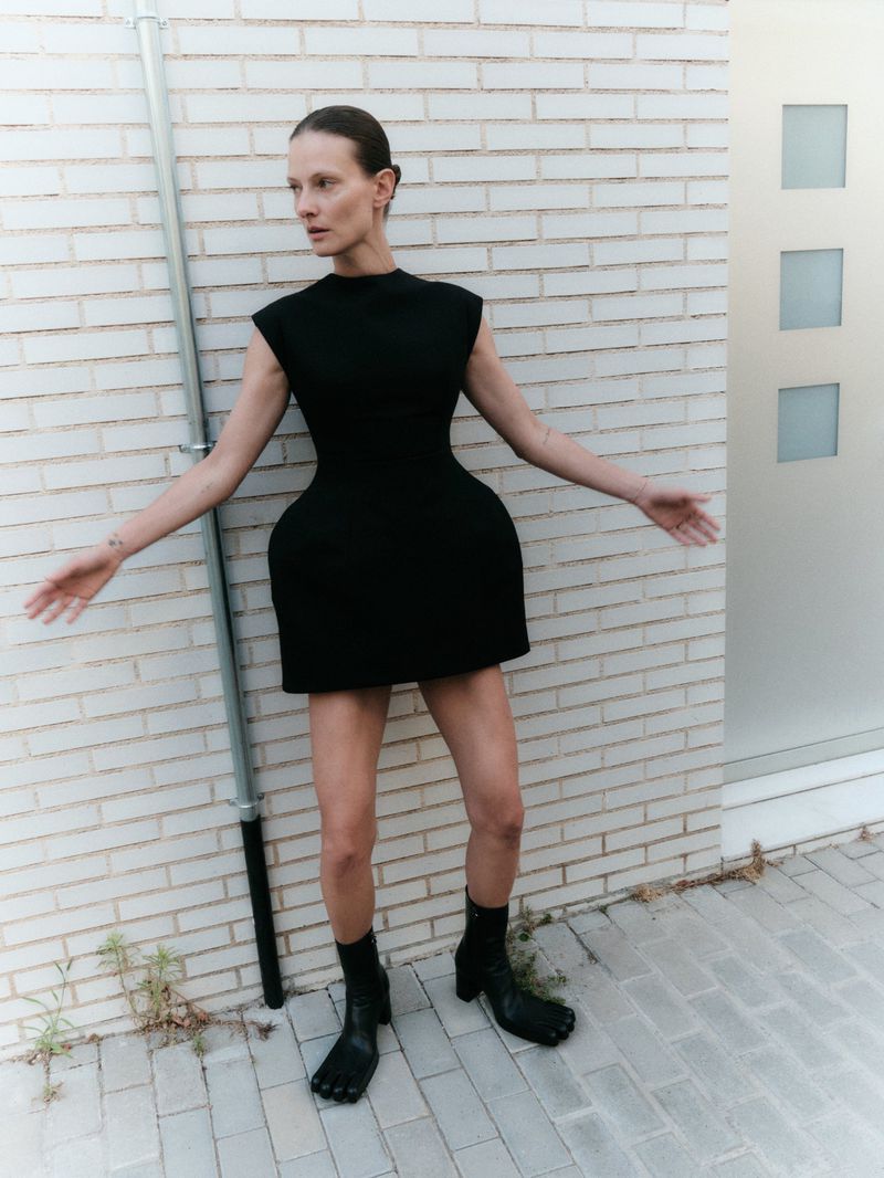 Model Tanya Ruban wearing a sculptural black sleeveless mini dress and black tabi boots against a pale brick wall.