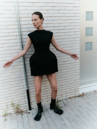 Model Tanya Ruban wearing a sculptural black sleeveless mini dress and black tabi boots against a pale brick wall.