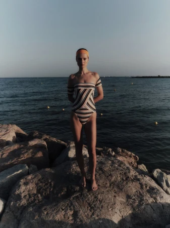 Tanya Ruban wearing a striped off-the-shoulder swimsuit and orange headband while standing on coastal rocks by the sea.