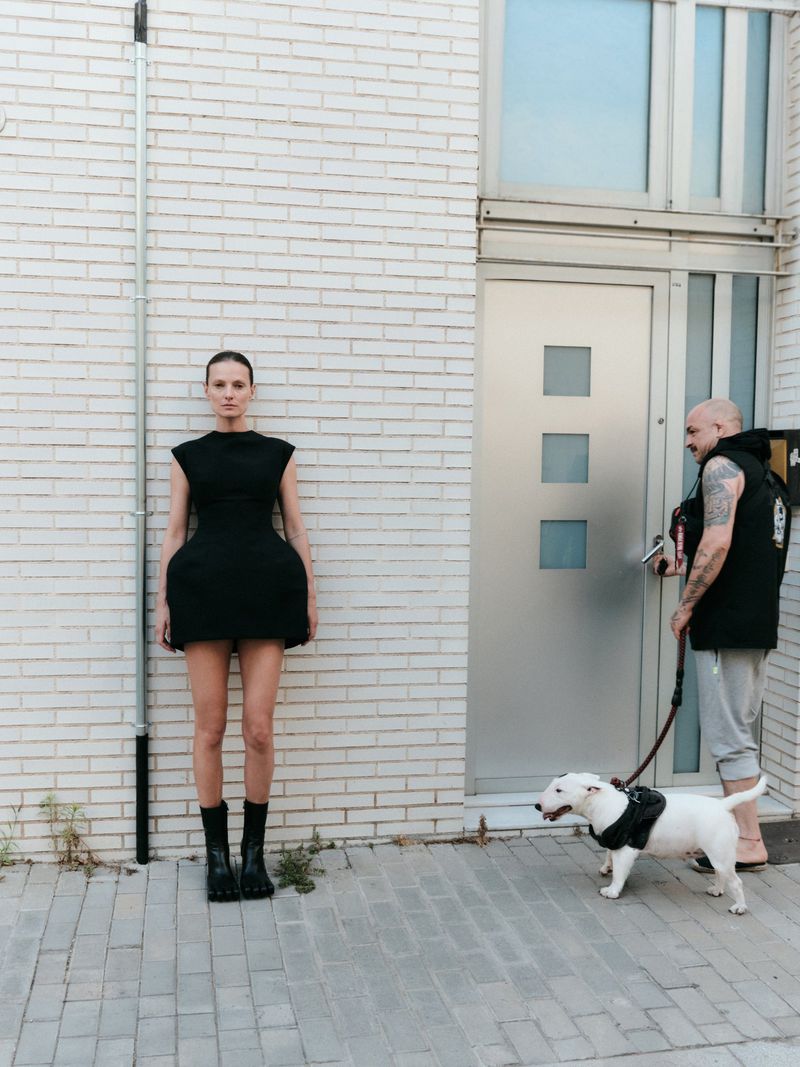 Model Tanya Ruban in a structured black mini dress standing against a white brick wall as a man walks a white bull terrier past a door.