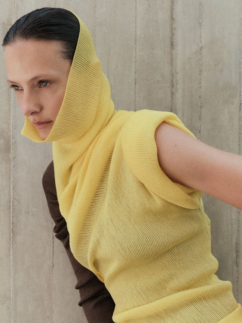 Model Tanya Ruban wearing a sleeveless yellow knit hood top and brown sleeve against a concrete background.