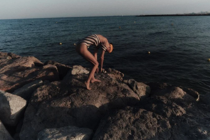 Model Tanya Ruban in a striped swimsuit bending forward on coastal rocks by the sea for Duel Magazine.